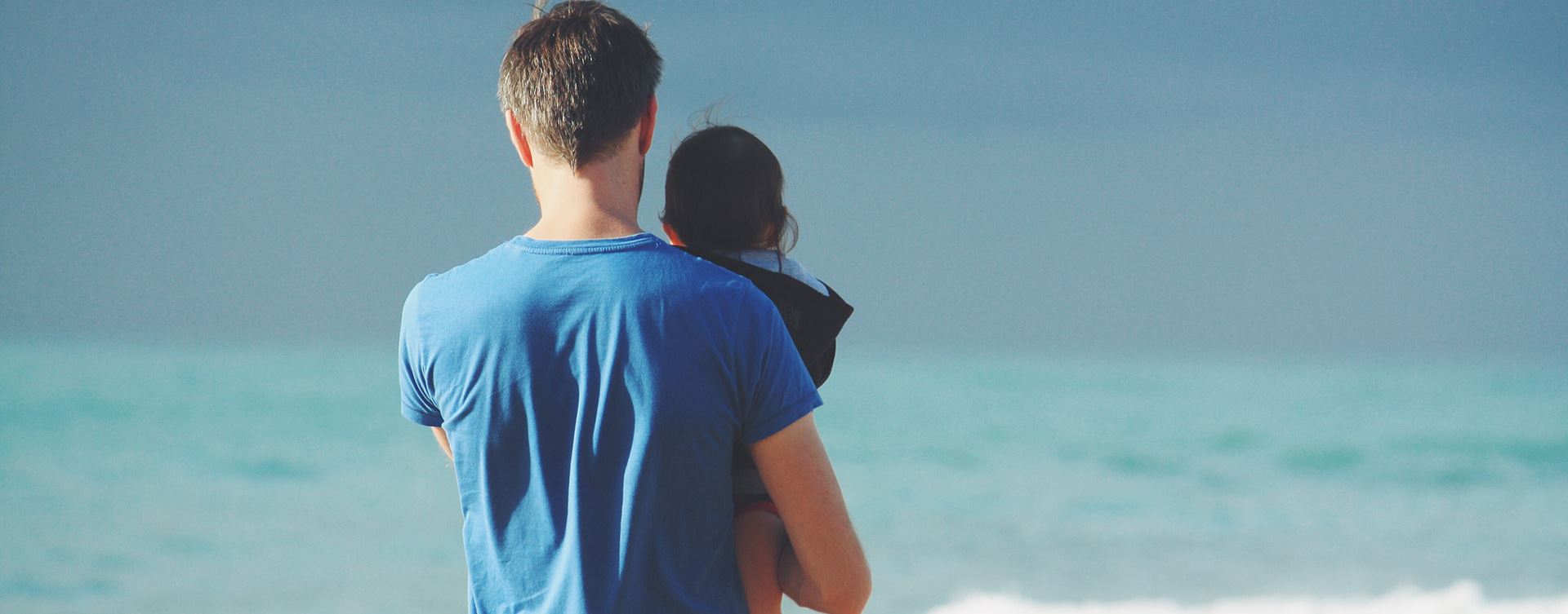 Dad holding young child on a beach facing away towards the sea