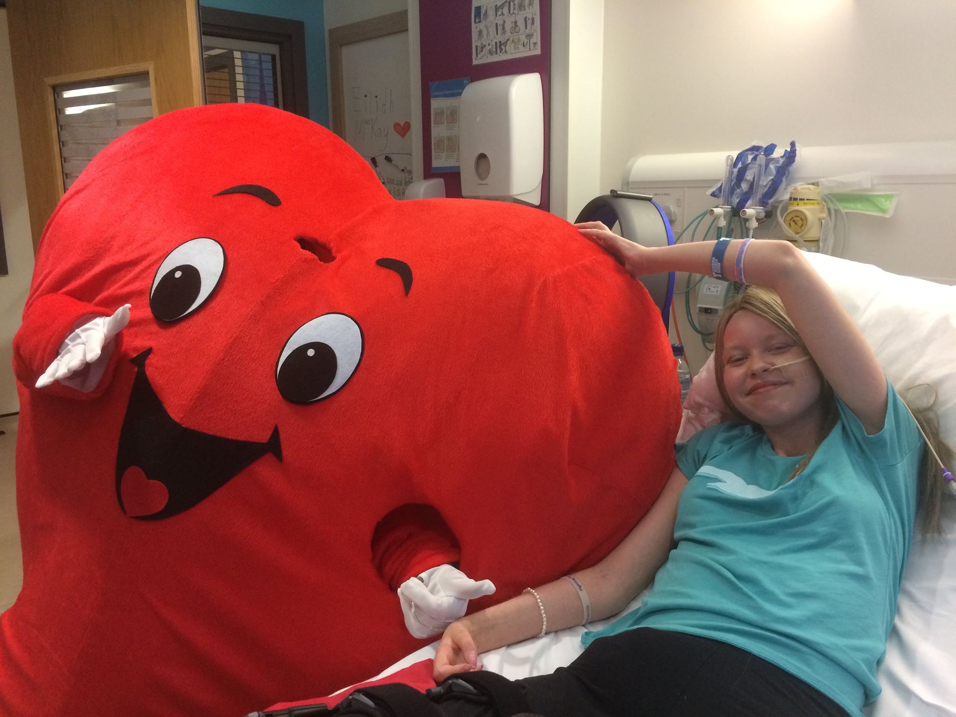 Eilidh in a hospital bed posing with a heart shaped mascot for Valentine's Day