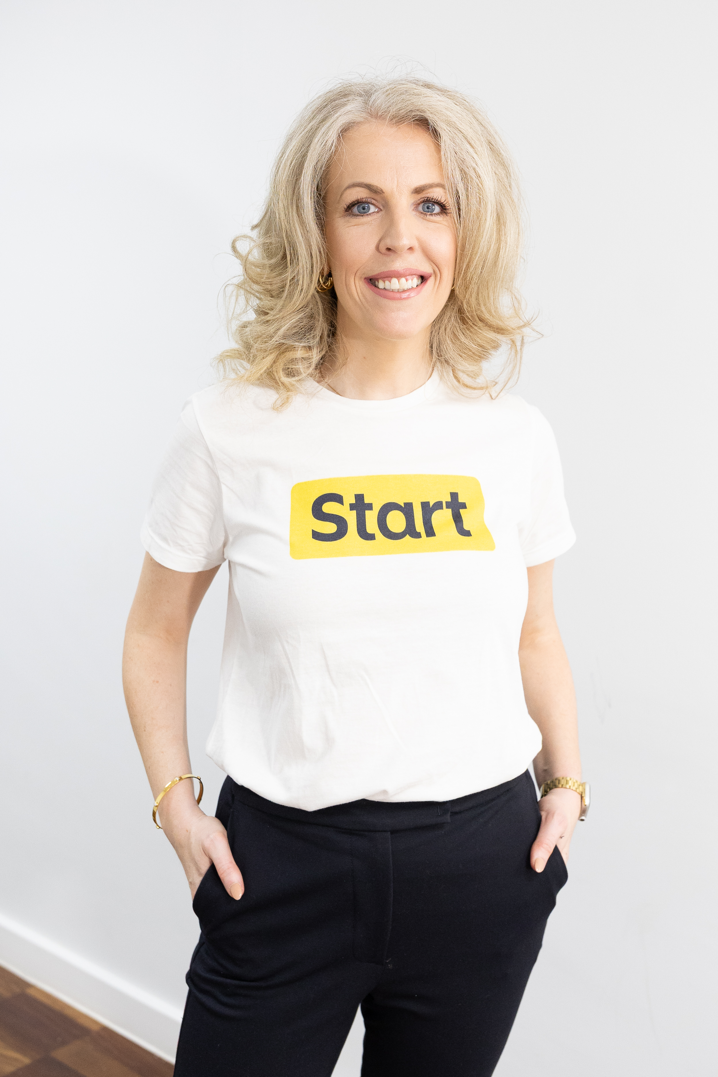 A woman with blonde hair smiles at the camera, wearing a white t-shirt with the word 'Start' written on it in navy and yellow