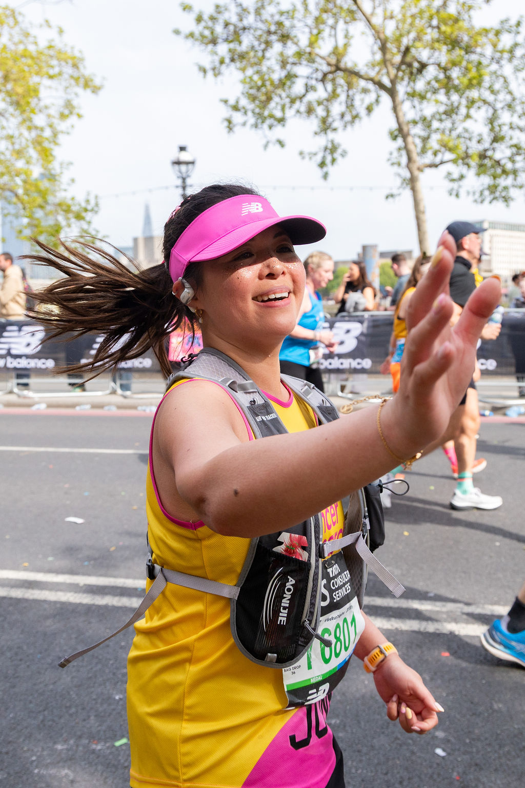 Runner in a yellow and pink vest and pink visor waves to the crowd during a marathon, hair blown sideways mid-stride, with other runners in the background.