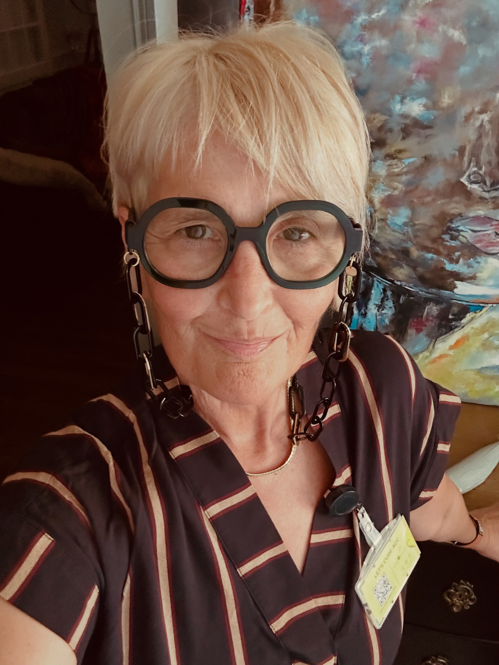 A selfie of a woman with short dark blonde hair and bold black framed glasses with a chunky chain. She is wearing a striped top and smiling gently