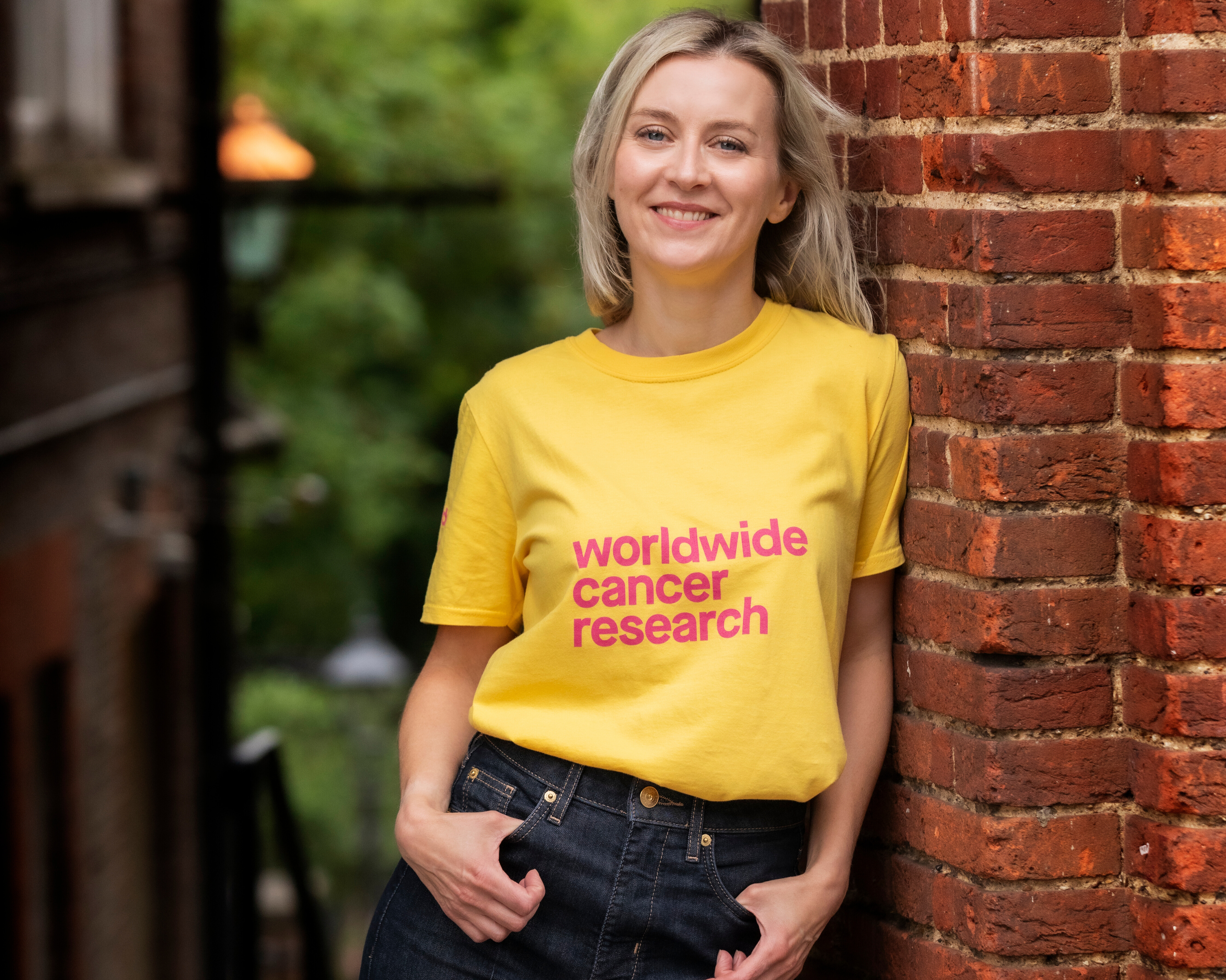 A photo of Dr Helen Rippon - a woman with shoulder length blonde hair and a big smile - looking directly at the camera, wearing a yellow Worldwide Cancer Research t-shirt and jeans