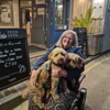 An image of a woman with grey hair and glasses, smiling, with two small dogs on her lap