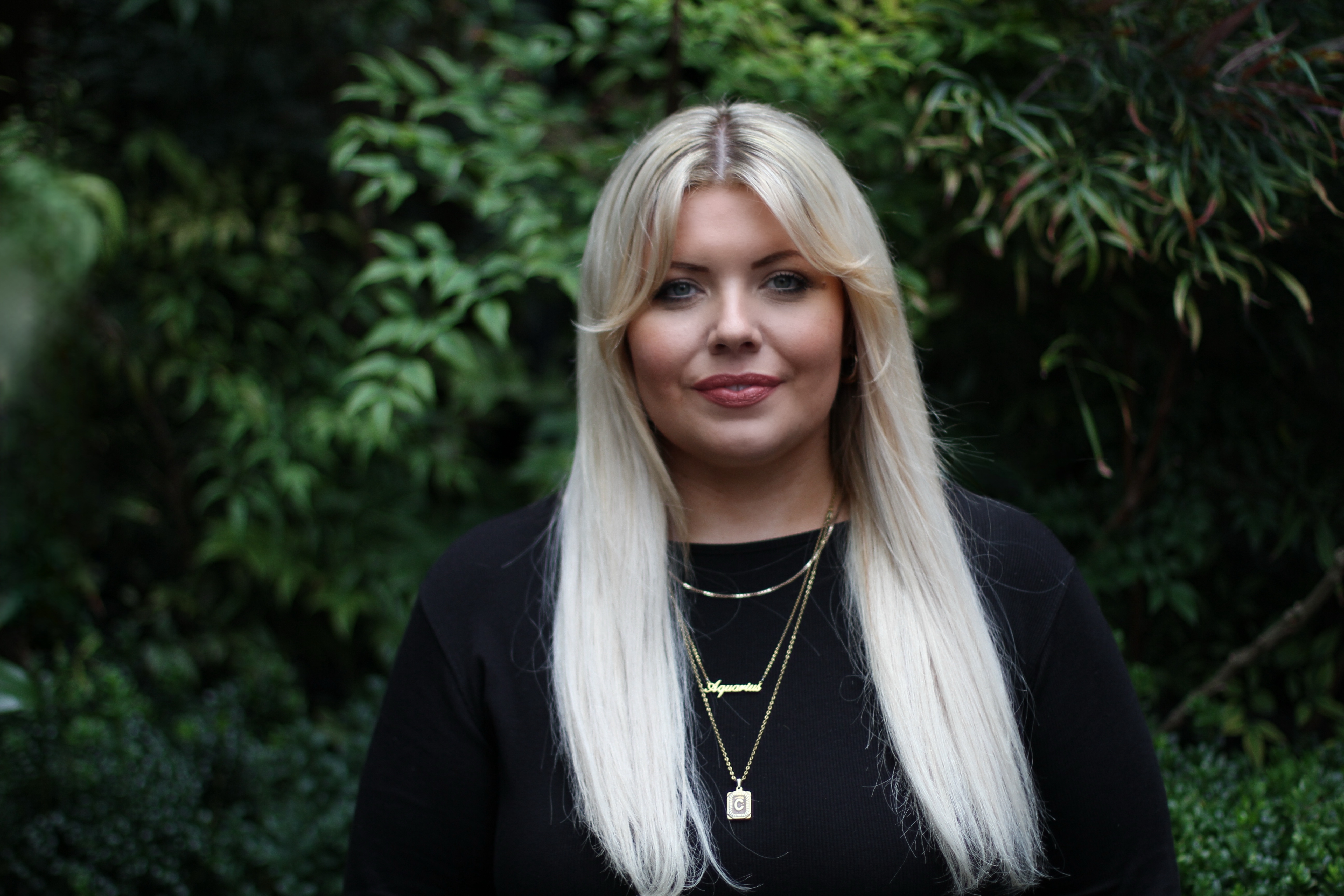A photo of a young woman with long blonde hair, standing in front of a tree and looking directly at the camera